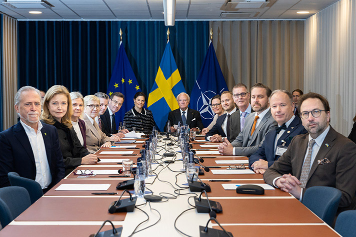 14 people sitting around large meeting room table, formal setting. In the background flags for the EU, Sweden and Nato.