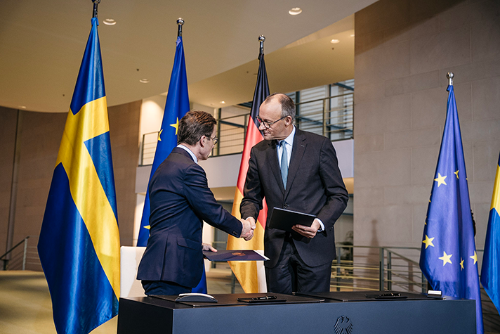 Ulf Kristersson and Friedrich Merz stand behind a desk indoors. They each hold a folder and shake hands. In the background the Swedish flag, the German flag and the EU flag.