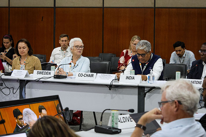 The two persons are ssitting at a conference table with particpants at both sides.