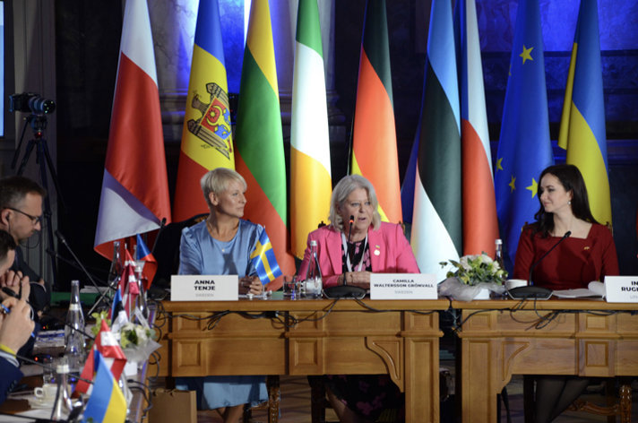 Ministers Anna Tenje and Camilla Waltersson Grönvall at a meeting table with flags in the background. Nameplates and microphones are placed on the table.