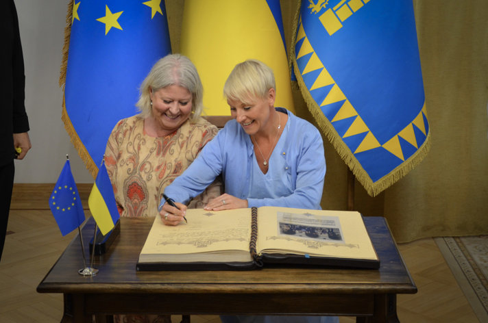Camilla Waltersson Grönvall and Anna Tenje sign a book. Behind them stand Ukrainian flags in blue and yellow. 