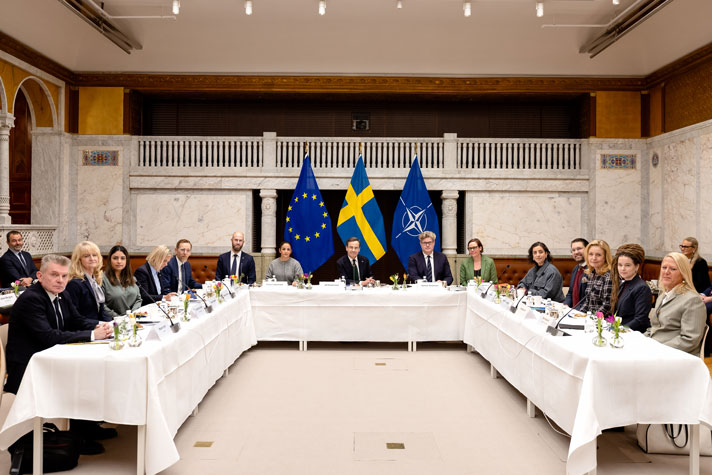 by party leaders/representatives of all parties represented in the Riksdag sitting at a u-shaped table.