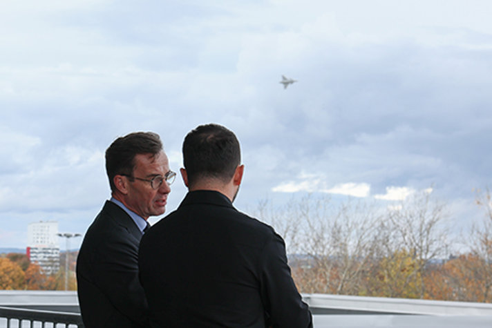 Outdoor image. Kristersson and Zelenskyy are watching as a JAS Gripen flies past.