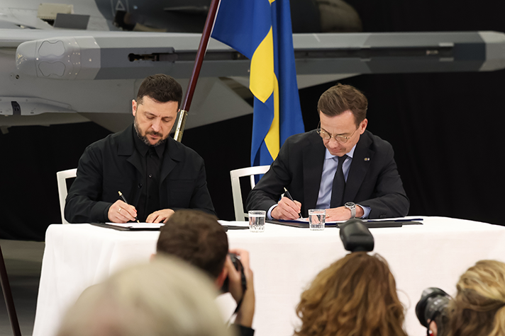 Volodymyr Zelensky and Ulf Kristersson sitting at a table, each signing a paper. Journalists are visible in the foreground.
