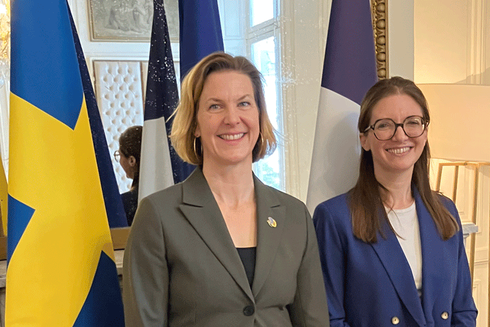 The Swedish and French Ministers for Gender Equality stands in front of the Swedish and French Flag. On the wall behind there is a mirror.