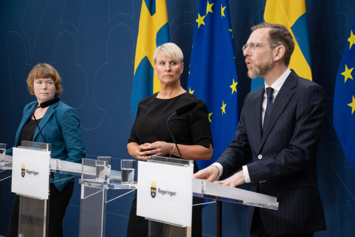 Minister for Older People and Social Security Anna Tenje, Minister for Social Affairs and Public Health Jakob Forssmed and Malin Danielsson, senior policy spokesperson for the Liberal Party stands in the Government's press room behind podiums. Behind them are Swedish flags and flags of the European Union. 