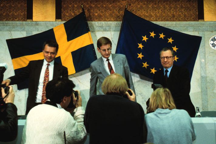 Ulf Dinkelspiel, Carl Bildt and Jaques Delors are standing in front of a table and some photographers. In the background there is a Swedish flag and a EU flag.