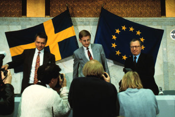 Ulf Dinkelspiel, Carl Bildt and Jaques Delors are standing in front of a table and some photographers. In the background there is a Swedish flag and a EU flag.